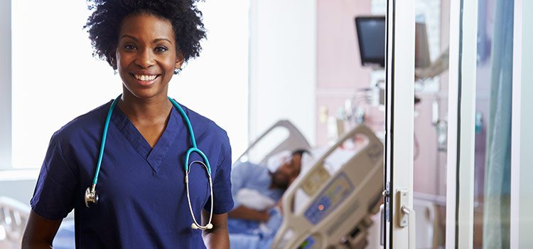 A nurse stands and smiles at the camera while a patient lies in bed in the background